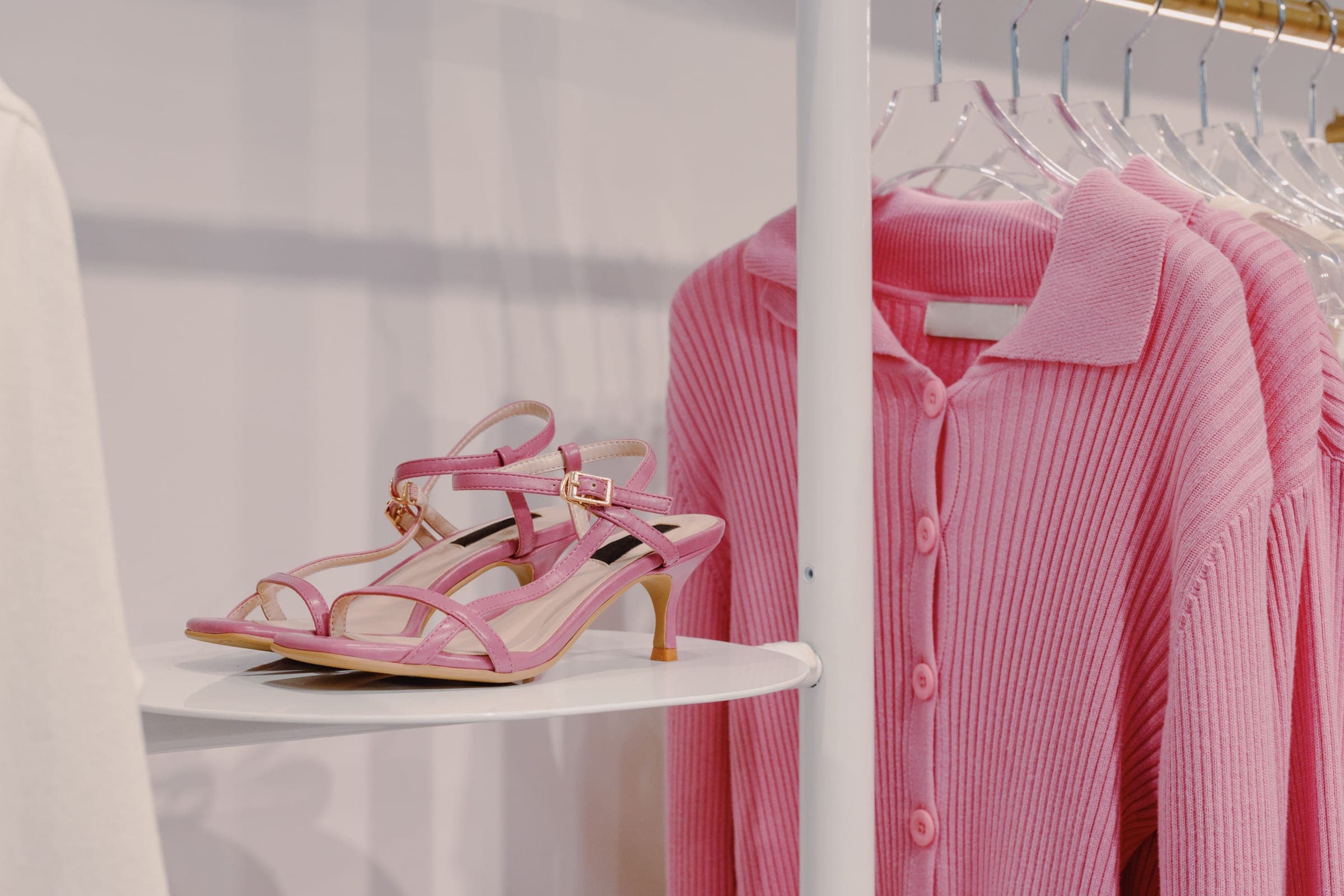 Pink strappy heeled sandals on a white shelf beside pink ribbed knit cardigans on a clothing rail