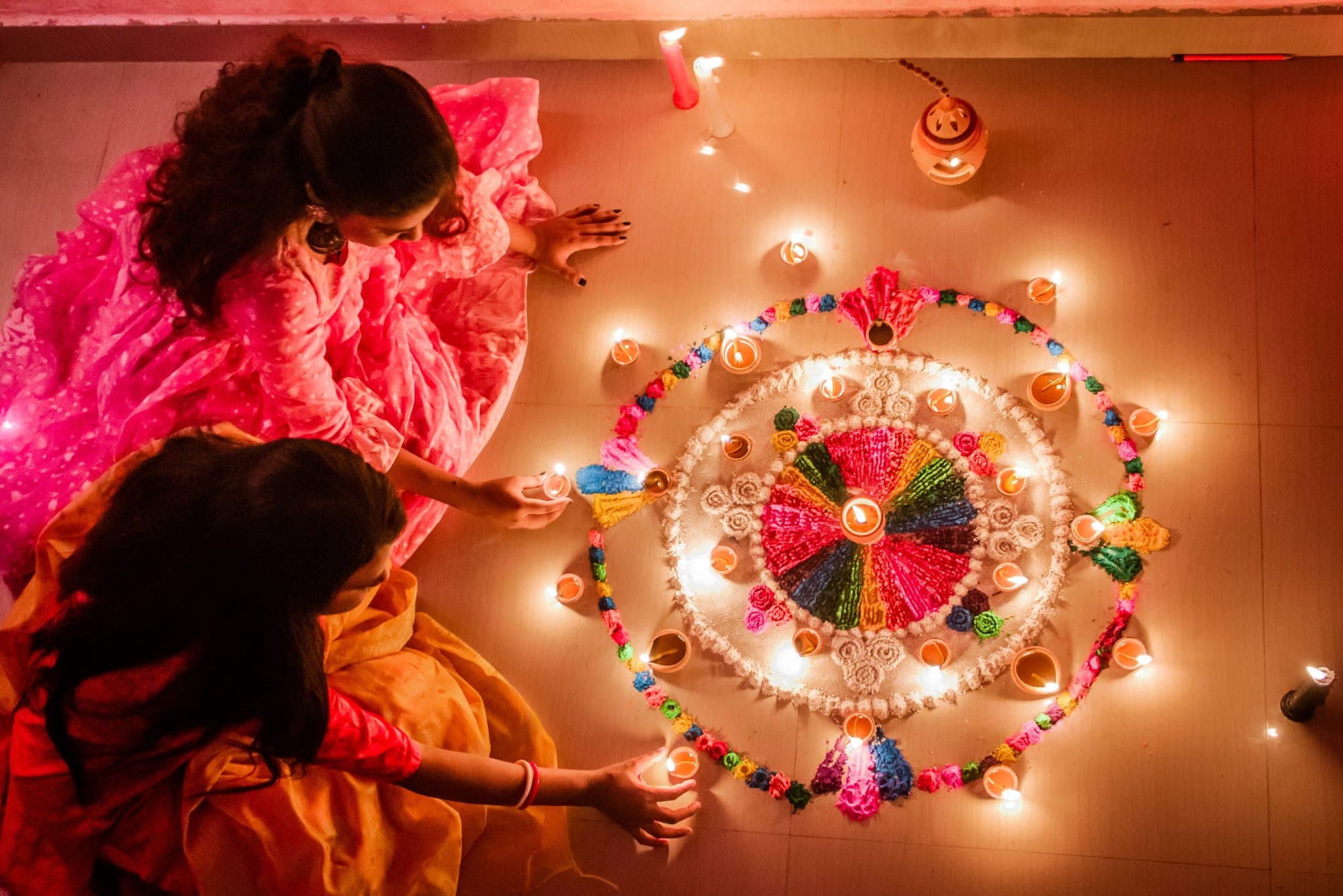 Two women in festive wear lighting diyas around a colourful rangoli, seen from above