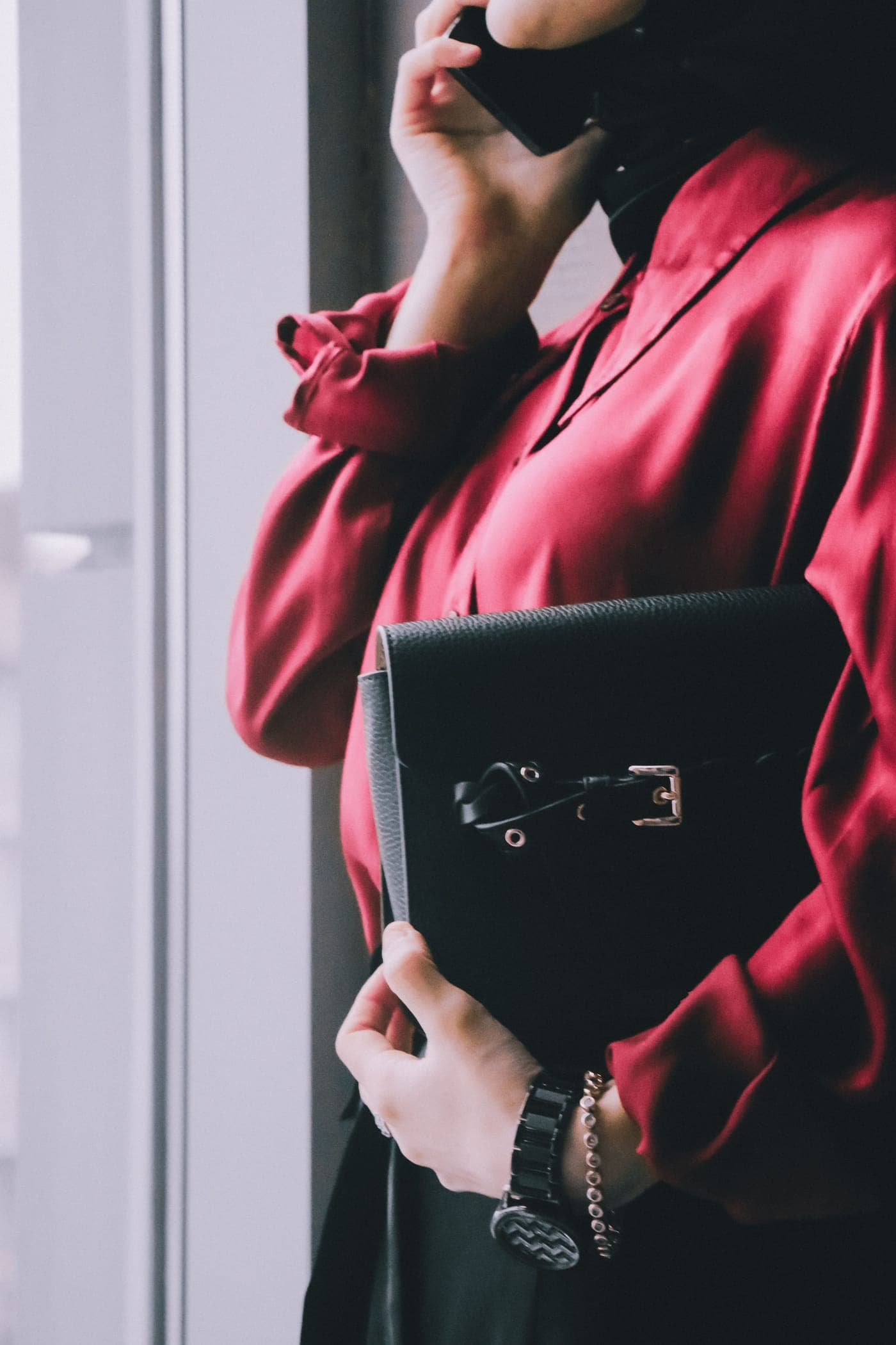 Woman in a red satin shirt holding a black leather bag while on a phone call by an office window