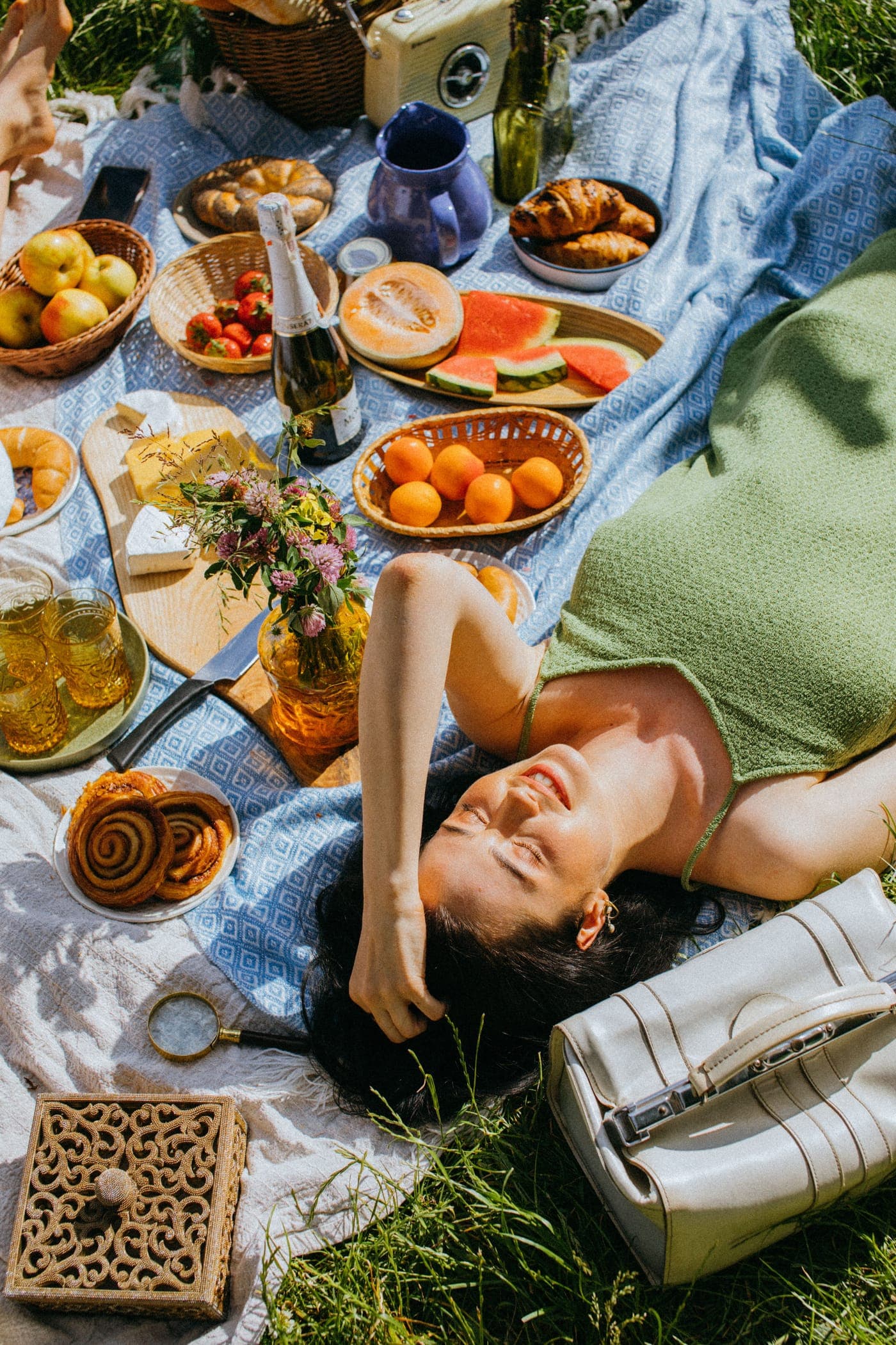 Woman in a green knit top relaxing on a picnic blanket with fruit, pastries and a white bag in the sun